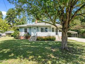 Single-story white house with a front porch, brick foundation, and a large tree in the yard. Concrete driveway extends to a carport on the right. Green lawn and shrubs surround the house, with trees in the background.