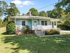 A single-story house with light blue siding, white trim, a front porch with steps, and a white door, surrounded by a green lawn, bushes, and trees under a partly cloudy sky.