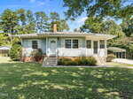 Charming single-story house at 608 15th Street, Butner, with light blue siding, white trim, and a brick foundation. Features a small porch, white front door, carport to the right, and is surrounded by trees and grass.