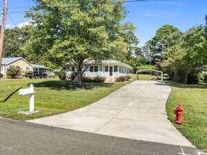 Single-story house with a large front yard, white mailbox, and red fire hydrant. A long concrete driveway leads to a covered parking area. Trees and grass surround the home, which has a front porch and multiple windows.