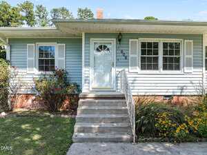 A light blue house with white shutters and trim, a glass-paneled front door, six concrete steps leading to the entrance, and landscaping with bushes and yellow flowers in front. House number 608 is by the door.