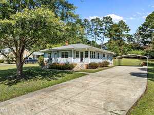 Single-story white house with a wide front porch and gray roof, set back from the street. A long concrete driveway leads to a metal carport, surrounded by green lawn, trees, and bushes under a clear blue sky.