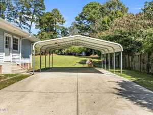 A metal carport with a peaked roof stands on a concrete driveway next to a blue house. The area is surrounded by trees and a wooden fence, with a grassy yard in the background under a clear sky.