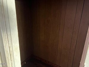 A small, empty closet or storage space with wood-paneled walls and a bare floor. The area is dimly lit, and the entrance is partially visible on the left side of the image.