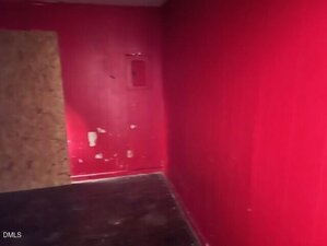 A small room with bright red walls, some scuff marks, a wooden board covering part of a doorway, a dark floor, and a red metal box mounted on the wall.
