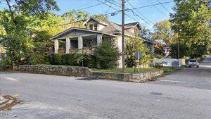 A two-story house with a covered front porch and dormer window sits on a corner lot at 1105 S Chestnut Street in Henderson. The yard features shrubs, a stone retaining wall, and faces a residential street with sidewalks in the foreground.