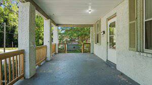 Covered front porch with a concrete floor, wooden railings, and white stucco exterior walls. There is a door and windows on the right side, with trees and greenery visible in the background.