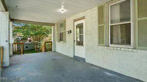 A covered porch with a concrete floor, a white door with a window, two green shuttered windows, and a wooden railing. The porch ceiling is white with a single light fixture. Trees are visible in the background.