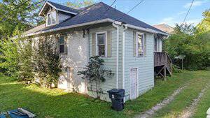 A small, light green house with gray roof shingles, white trim, and a side entrance. A trash bin sits by the house, and a grassy driveway runs alongside. There are shrubs and trees around the property.