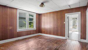 An empty room with wood-paneled walls, a wood floor with visible wear, a white ceiling with tiles, a ceiling light fixture, two windows, and an open doorway leading to a bright hallway.