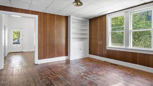 A room with wood-paneled walls, a worn wooden floor, a ceiling with white tiles, a window letting in daylight, and built-in white shelves with a cabinet. A doorway leads to another room in the background.