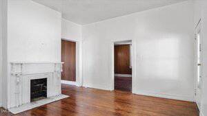 A room with white walls, a white fireplace on the left, and wood flooring. Two open doorways reveal adjacent rooms with similar wooden floors and brown doors. The room appears empty and well-lit.