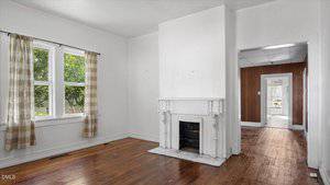 A room with white walls, a white decorative fireplace, hardwood floors, and a window with plaid curtains. An open doorway leads to another room with wood paneling and a view into a further space.