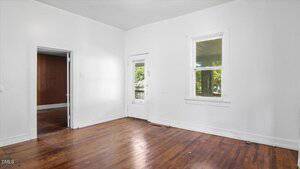 Empty room with white walls, two windows, and a wooden floor. A door with glass panels leads outside. An open doorway connects to another room with dark wood paneling. Natural light comes through the windows.
