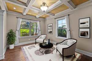 A small sitting room with two white armchairs, a round black coffee table, framed art on the wall, a large window, wood flooring, a potted plant, and a tray ceiling with a central light fixture.