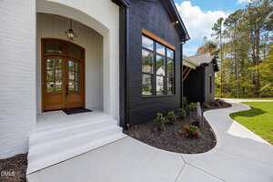 A modern house exterior features a white brick entrance with wooden double doors, adjacent to a black brick facade with large windows, landscaped shrubs, and a curved concrete walkway.