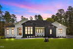 A modern single-story house at 3914 Sage Court in Wake Forest features a sleek black and white exterior, set on a green lawn among trees. Large windows glow from within as the sky turns pink and purple at sunset.