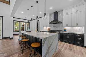 Modern kitchen with a large marble island, black and white cabinetry, bar stools, stainless steel appliances, and a herringbone tile backsplash. Pendant lights hang above the island, and wood flooring is visible.