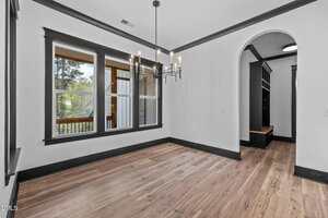 A bright, empty room with light wood floors, white walls, black trim, a modern black chandelier, large windows, and an arched doorway leading to another area with built-in shelving and a bench.