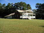 A single-story brick house with a metal roof and covered front porch sits on a large grassy lawn at 42 Tall Pines Drive, Henderson, bordered by trees in the background under a clear blue sky. Power lines run across the scene.