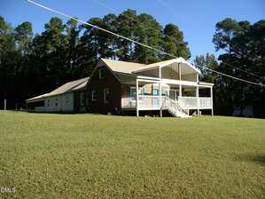 A single-story brick house with a metal roof and covered front porch sits on a large grassy lawn at 42 Tall Pines Drive, Henderson, bordered by trees in the background under a clear blue sky. Power lines run across the scene.