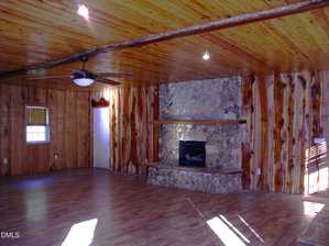 Large rustic living room with wood-paneled walls and ceiling, hardwood floors, a ceiling fan with light, and a stone fireplace with wooden mantel. Sunlight comes through a window, casting shadows on the floor.