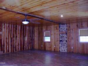 A large empty room with wood-paneled walls and ceiling, hardwood floors, two windows with blinds, a ceiling fan with a light, and a vertical stack of firewood built into one wall.