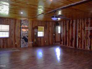 A spacious room features wood-paneled walls and a polished wood ceiling with exposed beams. There are several windows, a ceiling fan with lights, a stone fireplace, and a grandfather clock. The floor appears to be laminate wood.