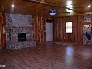 A spacious room with wood-paneled walls and ceiling, stone fireplace, ceiling fan, single door, a window with blinds, and laminate wood flooring. Stonework decor is also featured on the right wall.