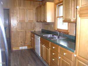 A kitchen with wood-paneled walls and cabinets, green countertop, double sink, dishwasher, and window above the sink; wood flooring and minimal decor.
