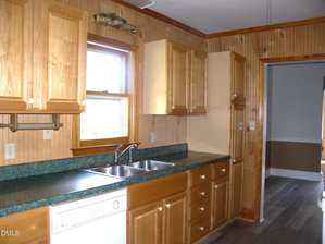 A kitchen with wood cabinets, green countertops, a double sink under a window, a dishwasher, and wood-paneled walls. The floor is dark with a visible adjoining room in the background.