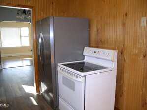A white electric stove and a stainless steel refrigerator are placed side by side against a wood-paneled wall in a kitchen. An open doorway leads to a bright room with windows and a ceiling light fixture.