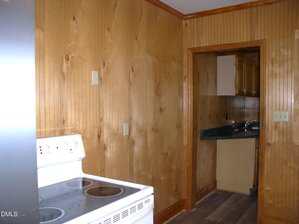 A kitchen with wood-paneled walls, an electric stove in the foreground, and a view into another room with matching paneling and green countertops. The floor is wood or laminate.
