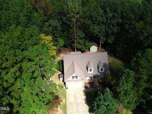 Aerial view of a two-story house with a gray roof and attached garage, surrounded by dense trees. A driveway leads to the garage, and a small shed is visible in the backyard near the edge of the woods.
