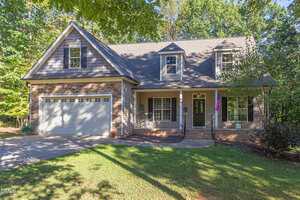 A two-story suburban house on 3627 Pine Needles Drive in Wake Forest with brick and siding exterior, an attached double garage, dormer windows, a covered front porch with an American flag, surrounded by trees and a front lawn.