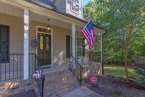 A front porch of a house with beige siding, black railing, brick steps, a hanging flower basket, an American flag, a CPI security sign, and a green yard with trees in the background.