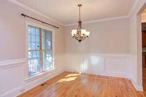 A bright dining room with light hardwood floors, beige walls with white wainscoting, a large window with grid panes, and a bronze chandelier hanging from the ceiling.