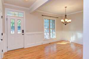 A bright, empty room with light hardwood floors, cream-colored walls with white wainscoting, a white front door with decorative glass, a large window, and a black metal chandelier.