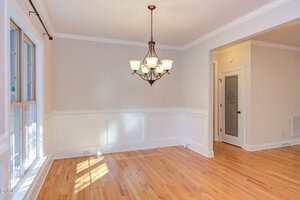 A dining room with hardwood floors, white wainscoting, beige walls, a window on the left, a six-light chandelier, and a door with frosted glass on the right side.