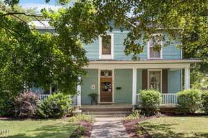 A charming two-story light blue house at 214 College Street in Oxford, featuring a large covered front porch, brown shutters, and white trim, surrounded by green shrubs and trees, with a concrete walkway leading to the front door.