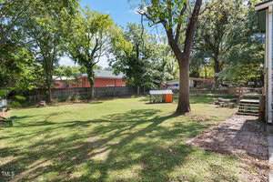A spacious backyard with grass, several mature trees, a wooden fence, a small orange shed, outdoor seating, and a brick patio area leading from steps on the right. Neighboring houses are visible through the trees.
