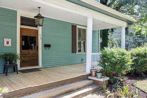 A wooden front porch with green siding, white trim, and a white column. A wooden door with glass, a house number "214," a black mailbox, and potted plants are visible. Shrubs and a small garden are beside the steps.
