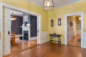 A hallway with yellow walls and wood floors leads to a room with dark walls, a fireplace, and a wooden dining table with chairs. The hallway features a small black table and wall art.