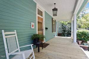 A wooden porch with light green siding features a white rocking chair, a potted plant, a black doormat, a wooden door with glass panel, a hanging lantern, and decorative house number on the wall. Trees are visible in the background.