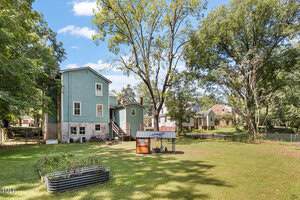 A large grassy backyard with a raised garden bed, a small metal-roofed chicken coop, and several mature trees. A light blue two-story house stands on the left, with neighboring houses visible in the background.