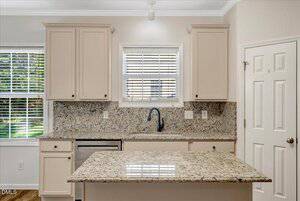 A modern kitchen with beige cabinets, granite countertops, a stainless steel dishwasher, a black faucet, a window with blinds above the sink, and an island in the center. Natural light enters from two windows.