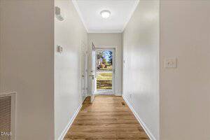 A hallway with light-colored walls and wood flooring leads to a closed front door with a glass storm door. Sunlight and greenery are visible through the glass door. The hallway has a ceiling light and wall switches.