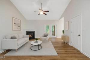 A modern living room with a light gray sofa and armchair, a round coffee table, a fireplace, wooden floors, a ceiling fan, and glass doors leading outside. Minimalist decor includes framed art and a sideboard.