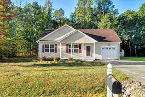 A single-story beige house with a red roof sits at 1220 E Middleton Drive E in Creedmoor. Featuring an attached garage, front porch, small shrubs along the facade, a green lawn, mailbox out front, and trees in the background.
