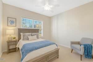 A neatly made bed with blue and white bedding sits against a wall beneath a window with blinds. A nightstand with a lamp and framed art is beside the bed. A gray armchair with a blue throw is in the corner.